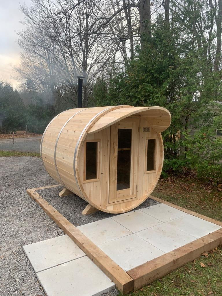 Wooden barrel-shaped sauna on a stone platform with trees in the background