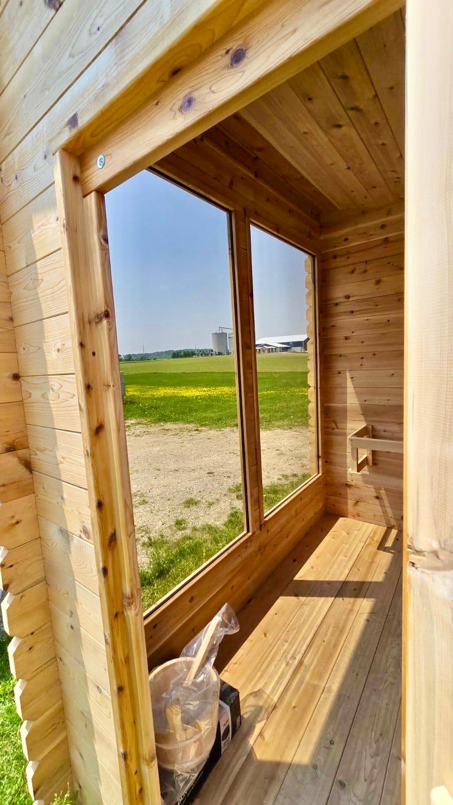 Wooden lean to sauna with a large window overlooking a field and blue sky