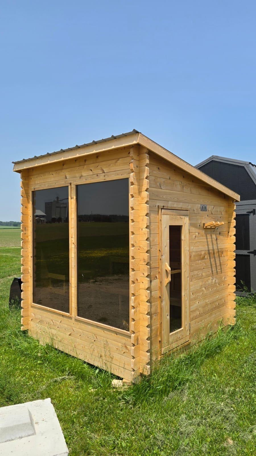 Wooden lean to sauna with large windows on a grassy area with a clear blue sky.