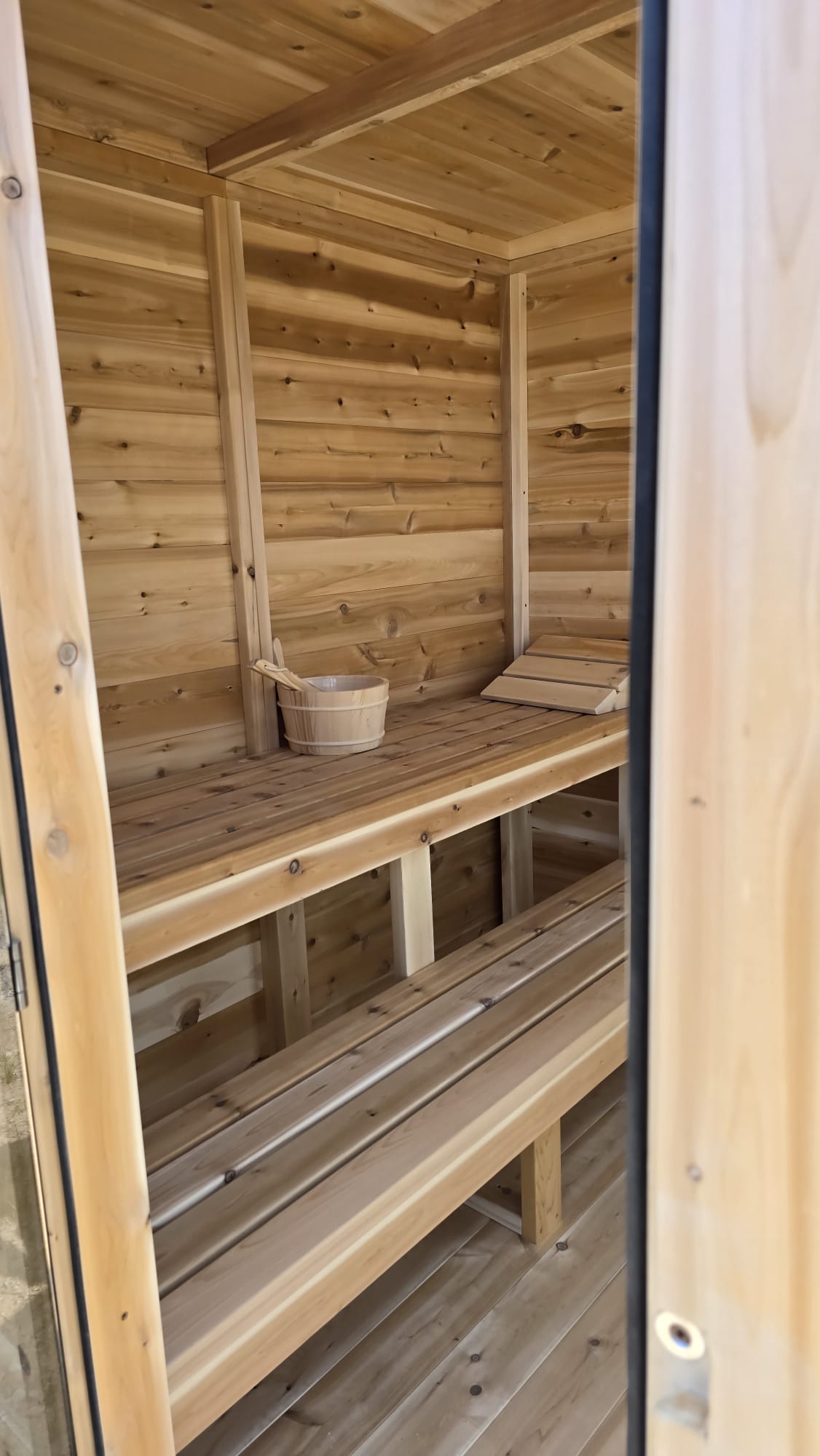 Wooden sauna with benches and a bucket inside with glass door