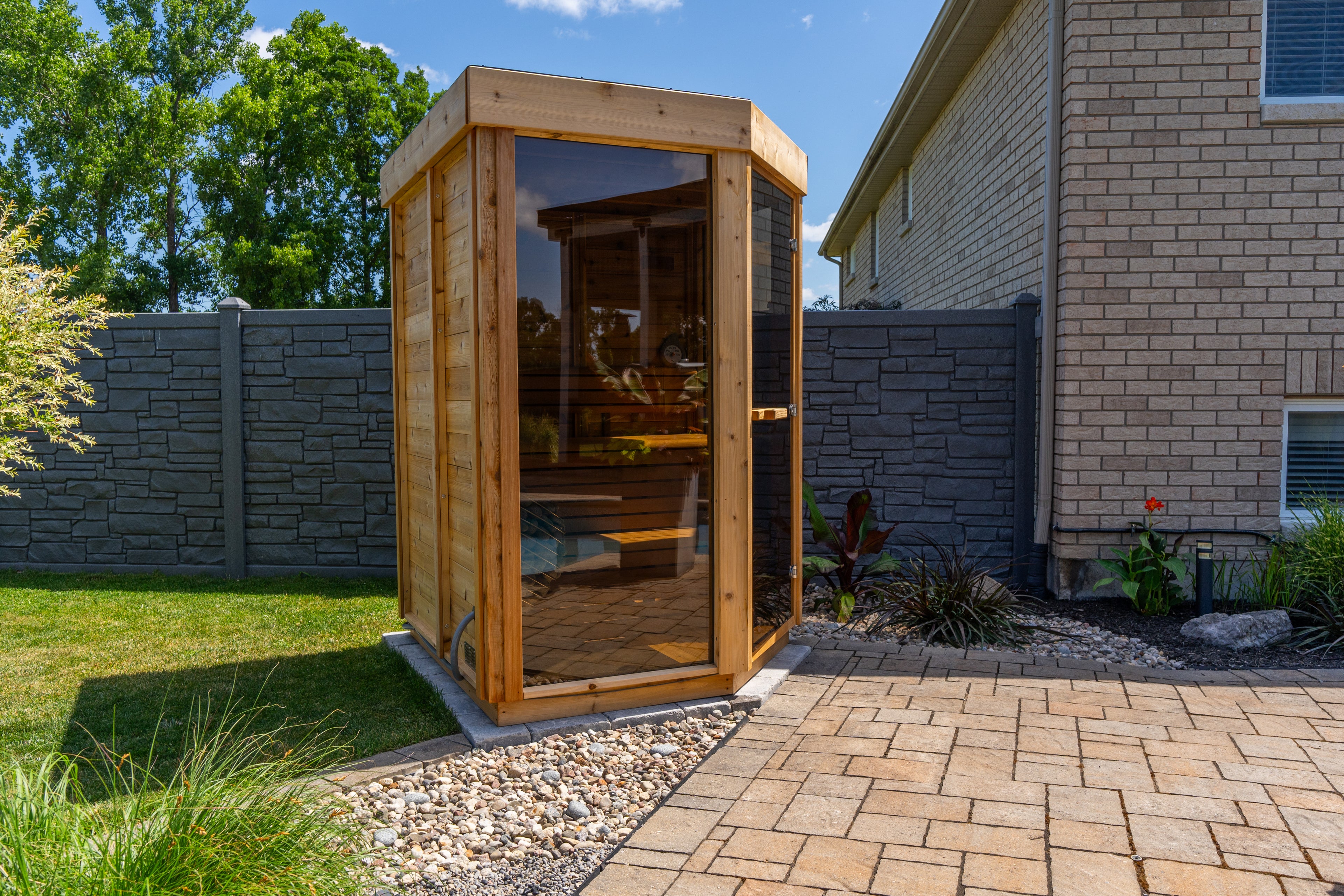 Wooden outdoor trinity sauna in a backyard setting with a brick patio and grass.