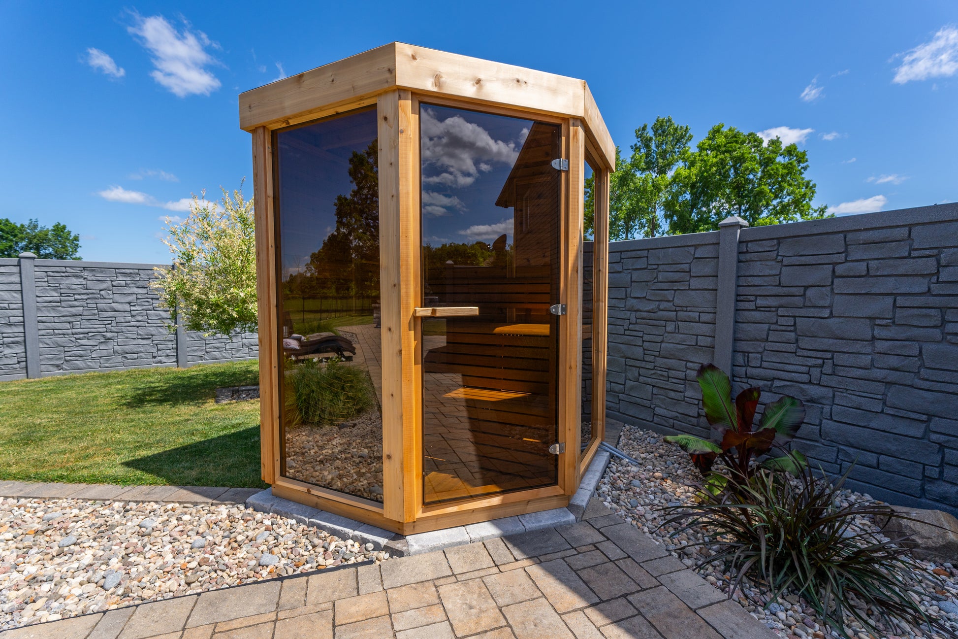 Wooden outdoor trinity sauna cabin with glass doors in a garden setting.