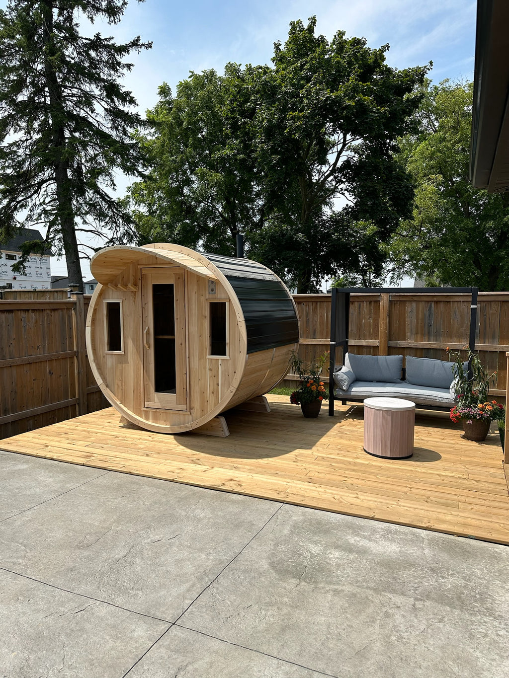 Wooden barrel sauna on a wooden deck with trees and a house in the background