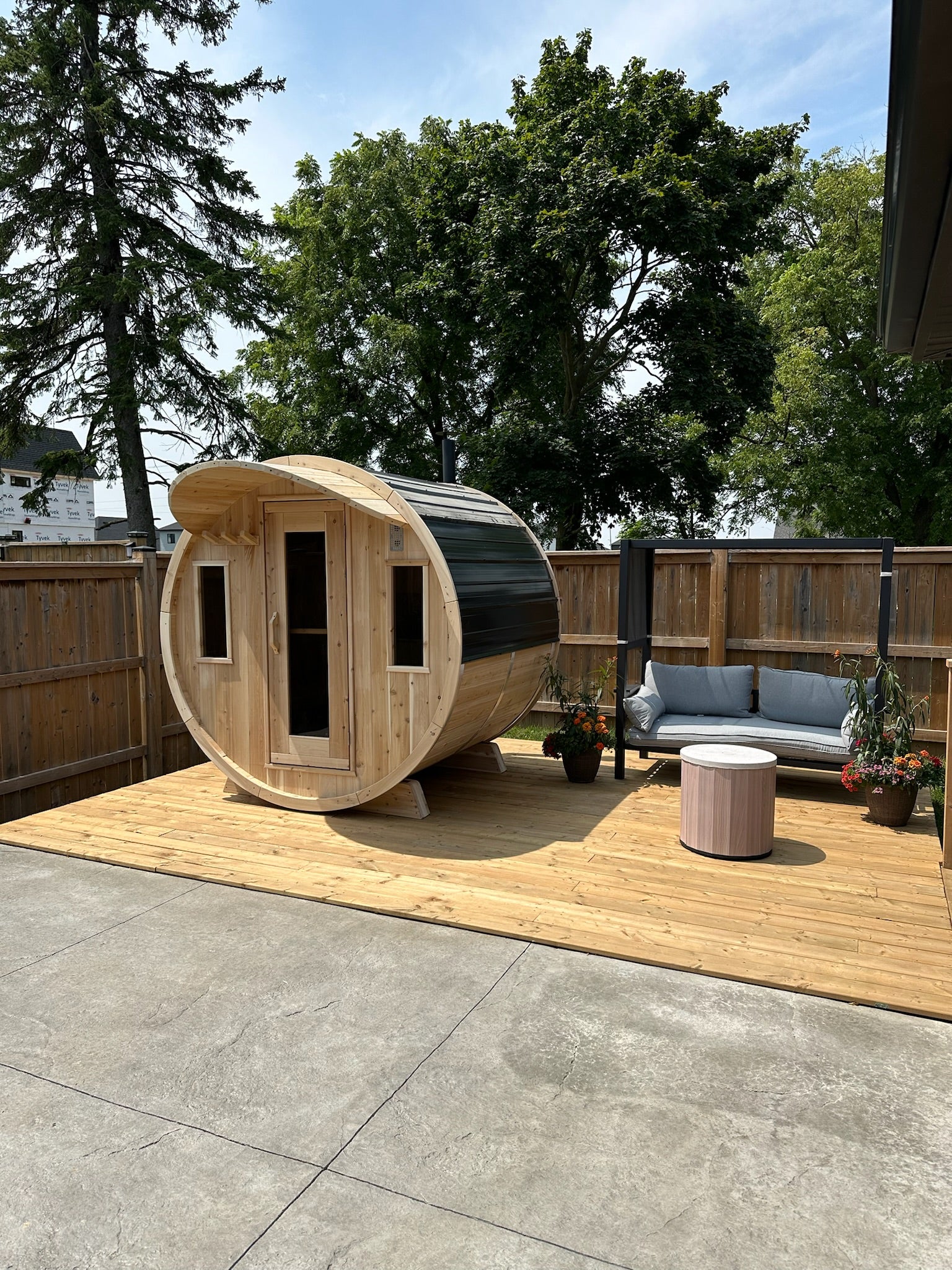 Wooden barrel sauna on a wooden deck with trees and a house in the background