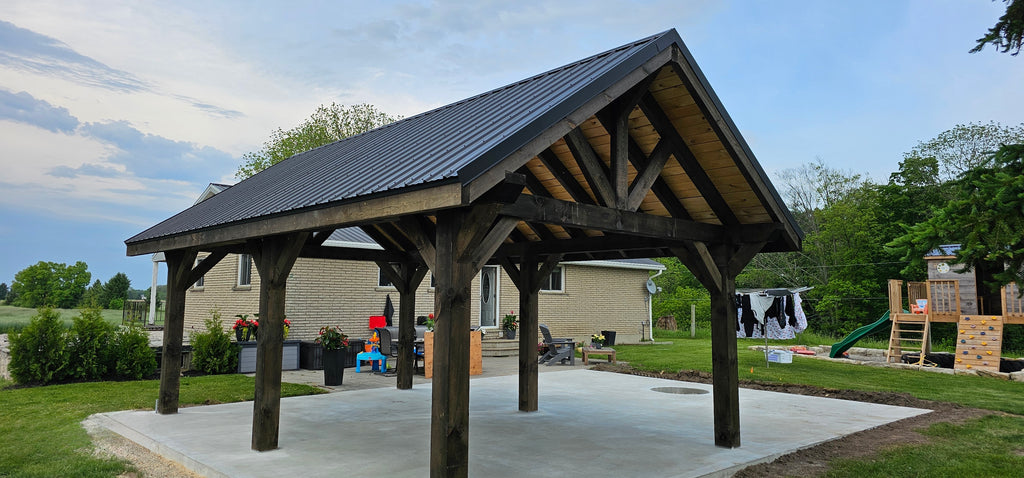 Wooden pavilion with a metal roof on a concrete slab, surrounded by greenery and outdoor furniture.