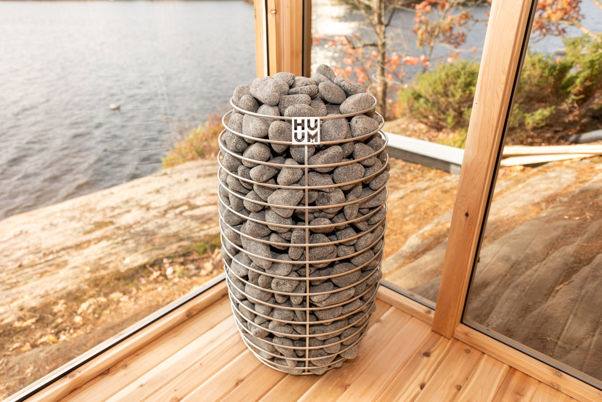sauna heater filled with stones in a sauna by a window with a view of water and trees.