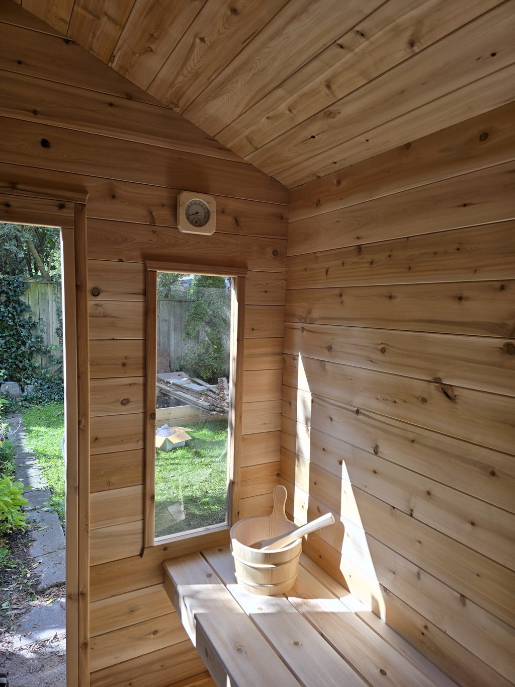 Wooden sauna with a bucket and bench, surrounded by greenery outside.