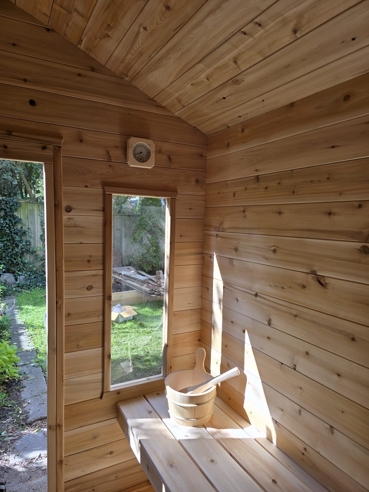 Wooden sauna with a bucket and bench, surrounded by greenery outside.